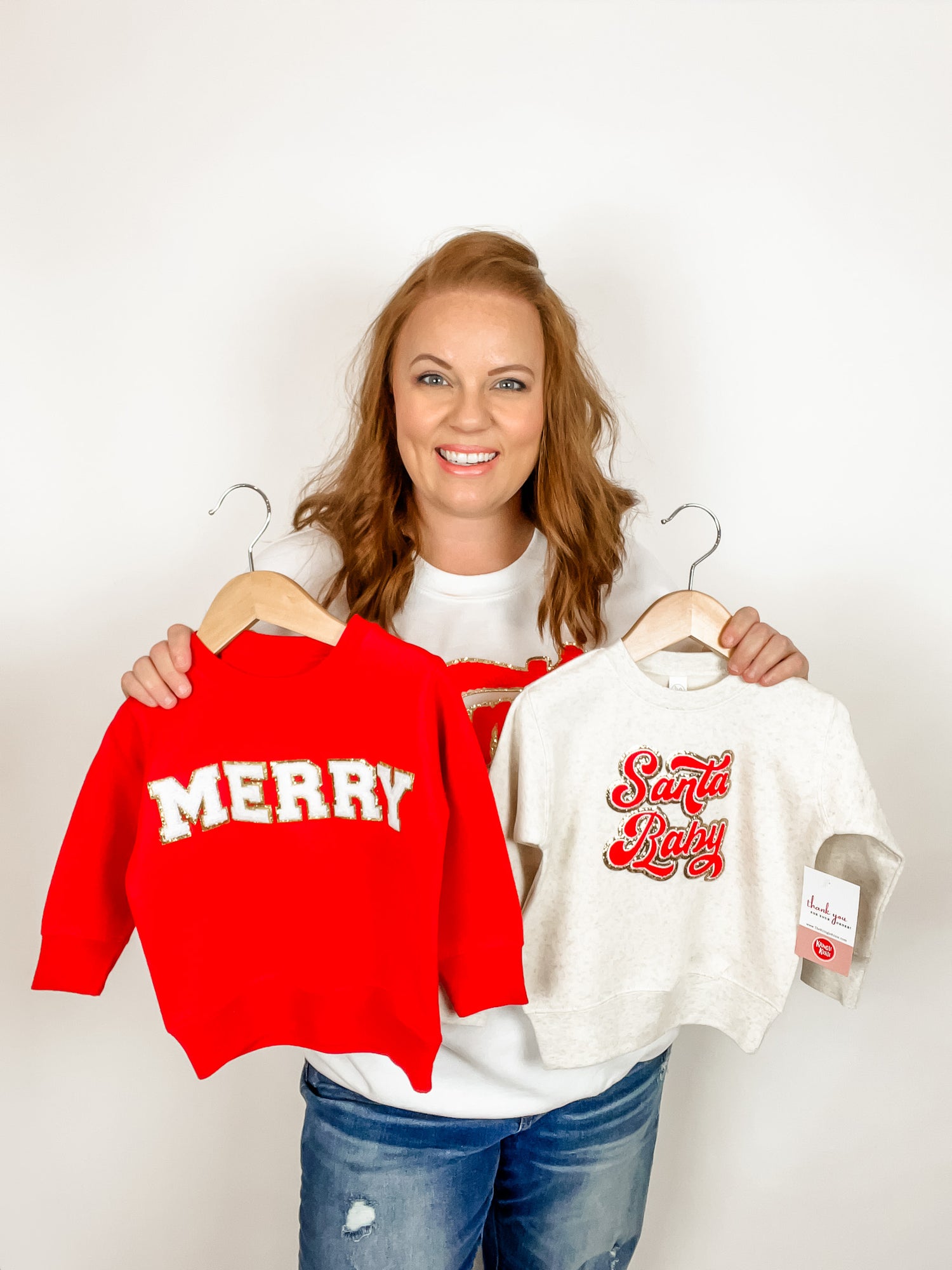 Abby Robertson, owner of the Kringle Krate Boutique holds two toddler sweatshirts in red and beige that feature chenille patches reading "Merry" and "Santa Baby."